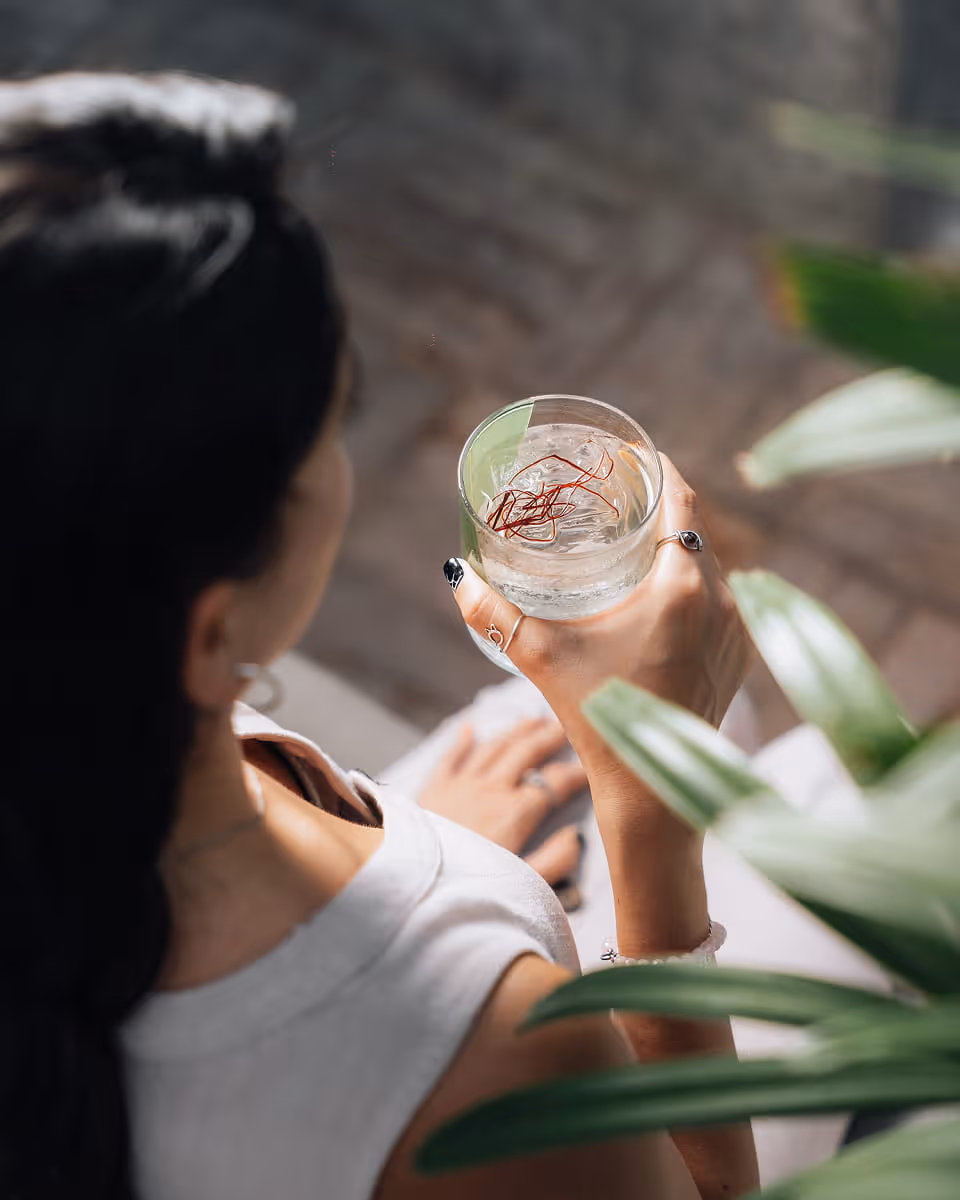 A woman holding a clear cocktail garnished with saffron threads and a lime wedge while relaxing near tropical plants