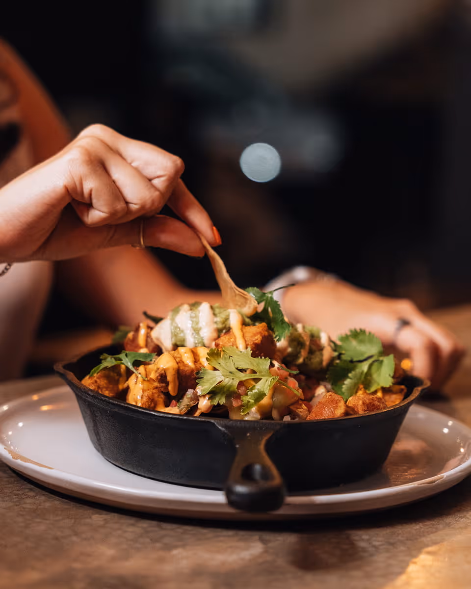A hand pulling a loaded nacho chip from a cast iron skillet of nachos topped with cilantro, salsa, and drizzled sauces