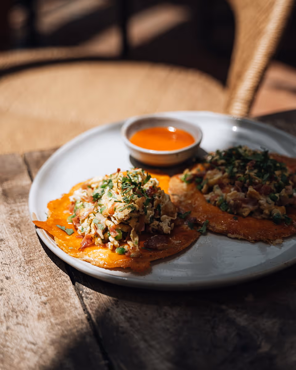 Crispy tostadas topped with scrambled eggs, bacon, cilantro, and a side of orange habanero salsa on a rustic wooden table