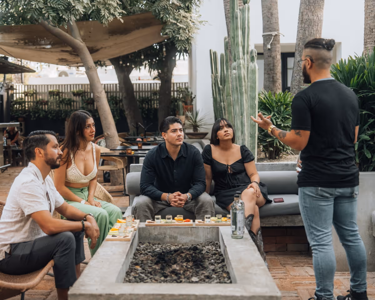 A guide leads a mezcal tasting experience for a group of four guests seated around an outdoor fire pit with cacti nearby.