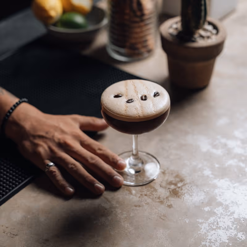 A bartender presents an espresso martini cocktail garnished with three coffee beans in a coupe glass on a marble bar counter.