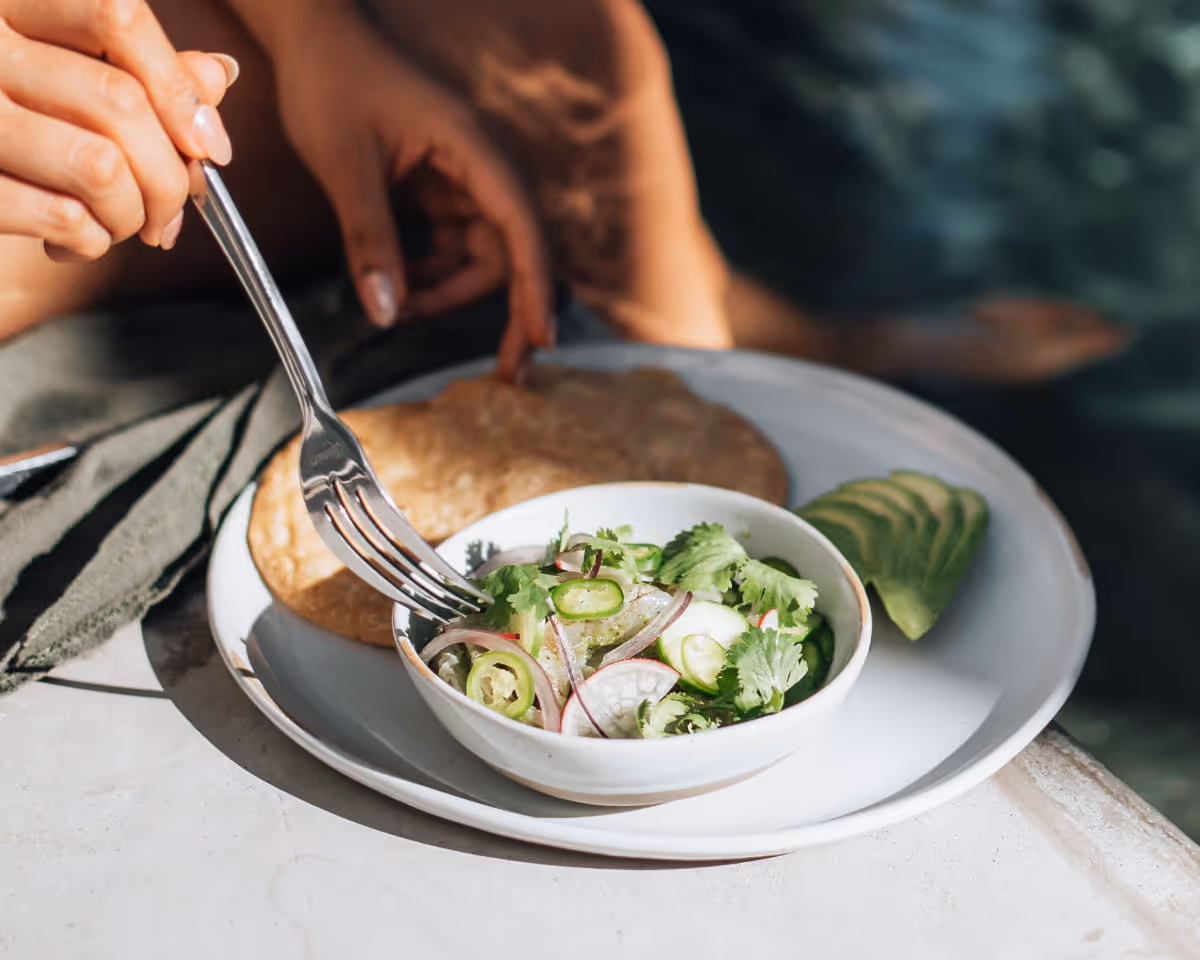 A person uses a fork to eat fresh ceviche with cilantro, radish, and jalapeño, served with tortilla and sliced avocado.