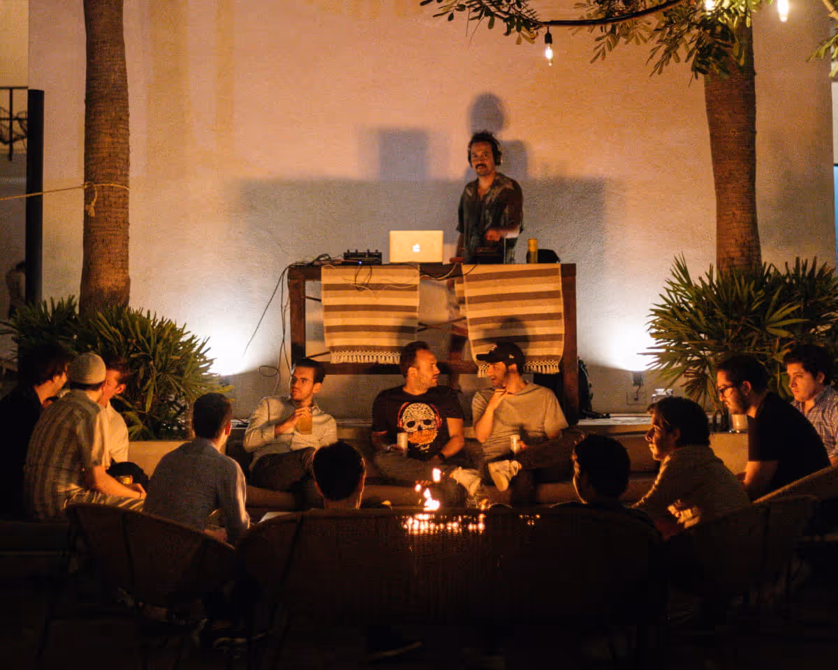 A DJ plays music at an outdoor nighttime gathering as guests sit around a fire pit in a tropical courtyard setting.