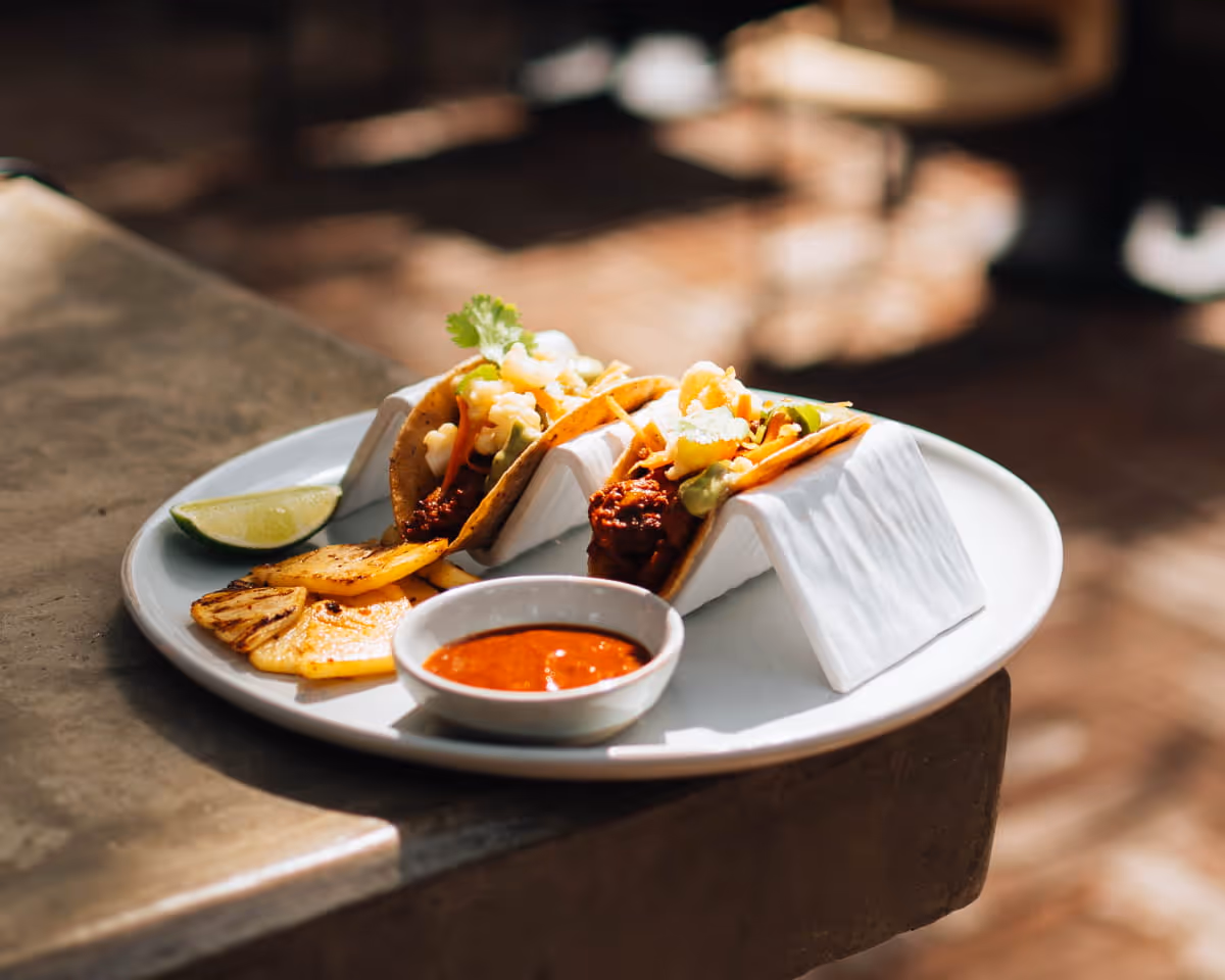 Three gourmet tacos served on a white plate with grilled pineapple, lime wedge, and red salsa in warm sunlight