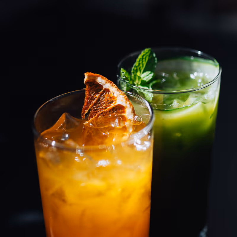 Two tropical cocktails on ice — an orange citrus drink with dried orange garnish and a green mint cucumber cooler