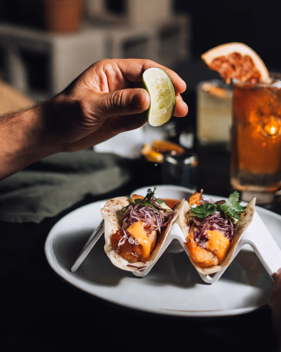 A hand squeezing a lime wedge over two fish tacos topped with red cabbage slaw and chipotle sauce on a white plate