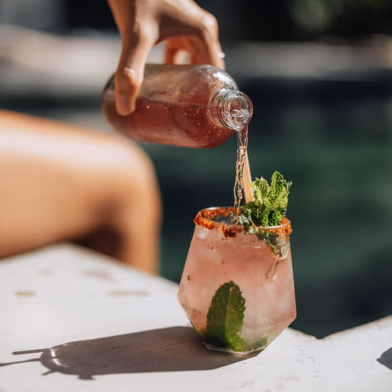 A hand pouring a pink cocktail from a bottle into a garnished glass with mint and chili rim by the poolside