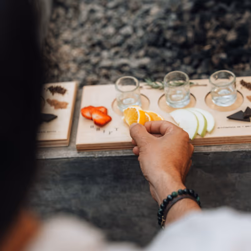 Hand reaching for an orange slice on a wooden tasting board with shot glasses, strawberries, apple slices, and chocolate.