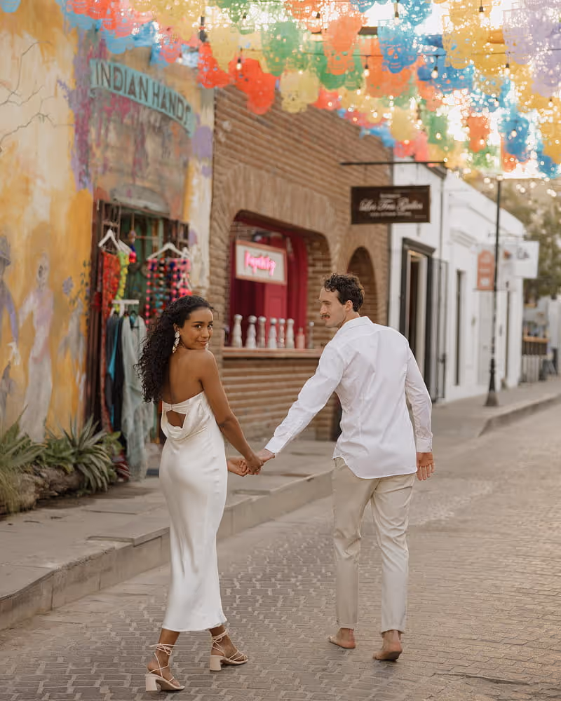 Newlywed couple walking hand-in-hand through a colorful Mexican street decorated with papel picado banners overhead