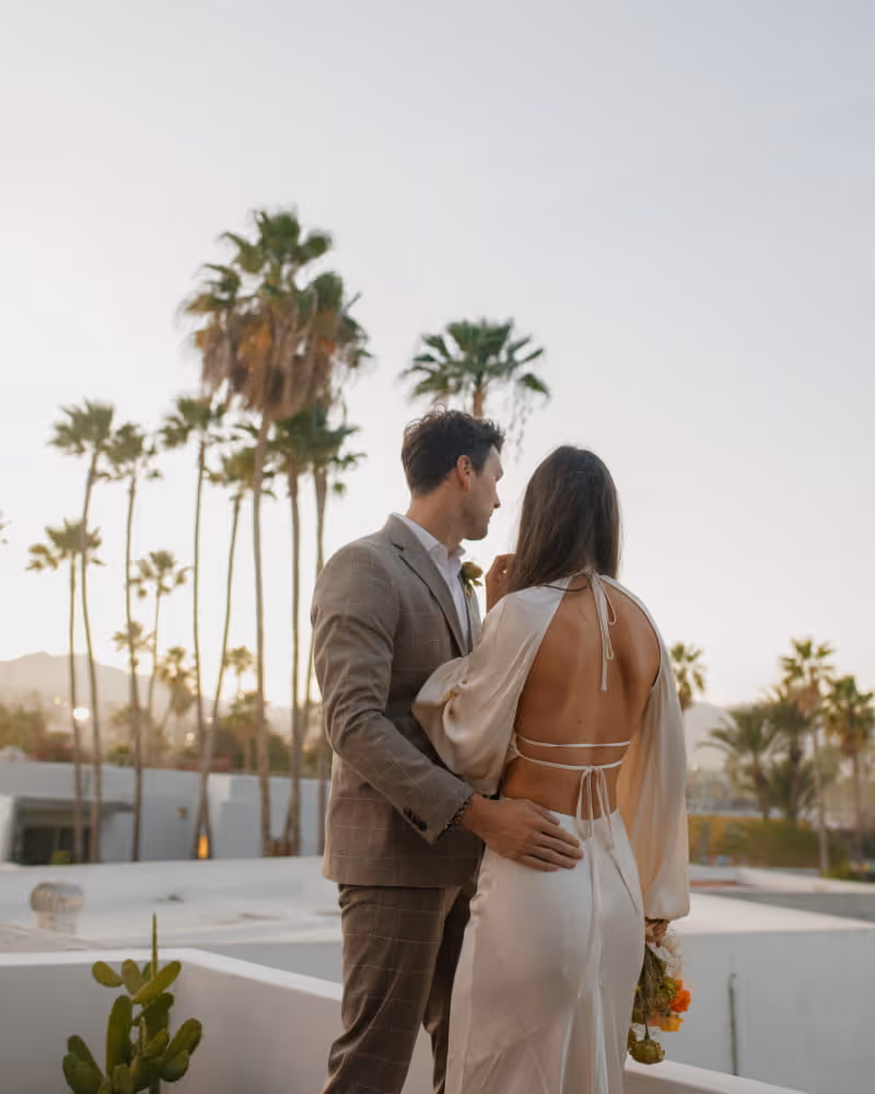 Couple embracing on a rooftop at golden hour with palm trees and desert mountains in the background during their wedding