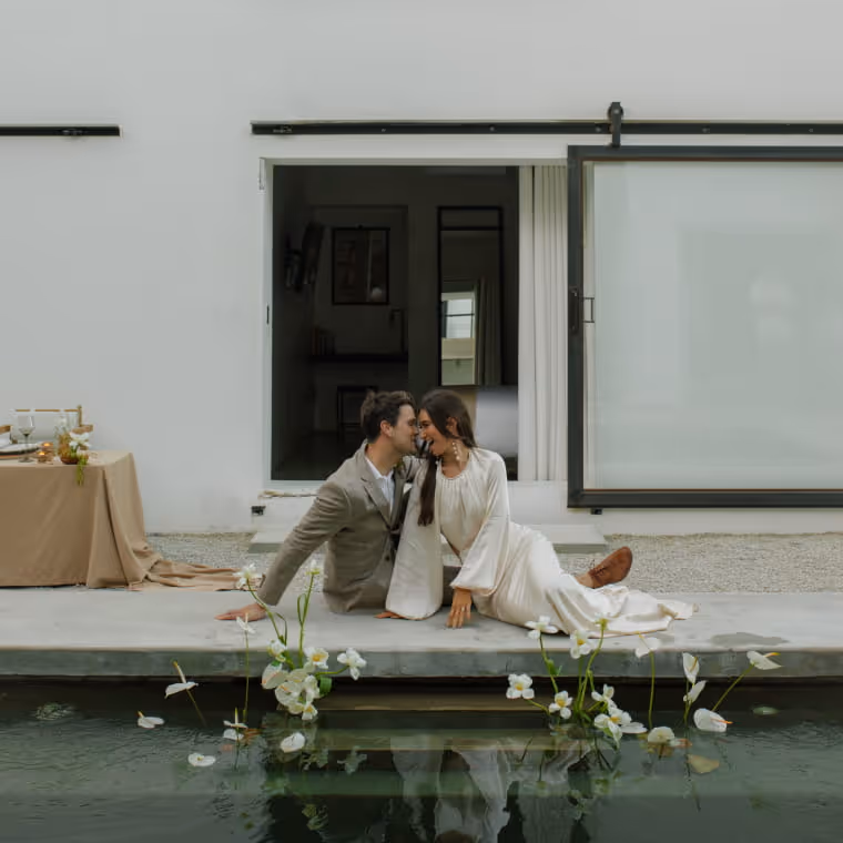 Couple sharing a kiss poolside beside a candlelit dinner table with white flowers floating in the water