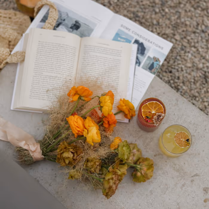Flat lay of orange and yellow wedding bouquet resting on open books beside citrus cocktails on a stone table