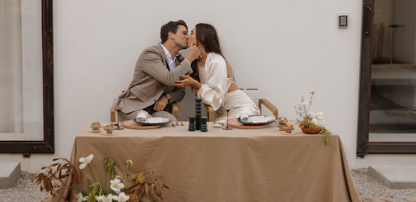Newlywed couple sharing a kiss at their intimate sweetheart table decorated with earth-toned linens, candles, and ceramic