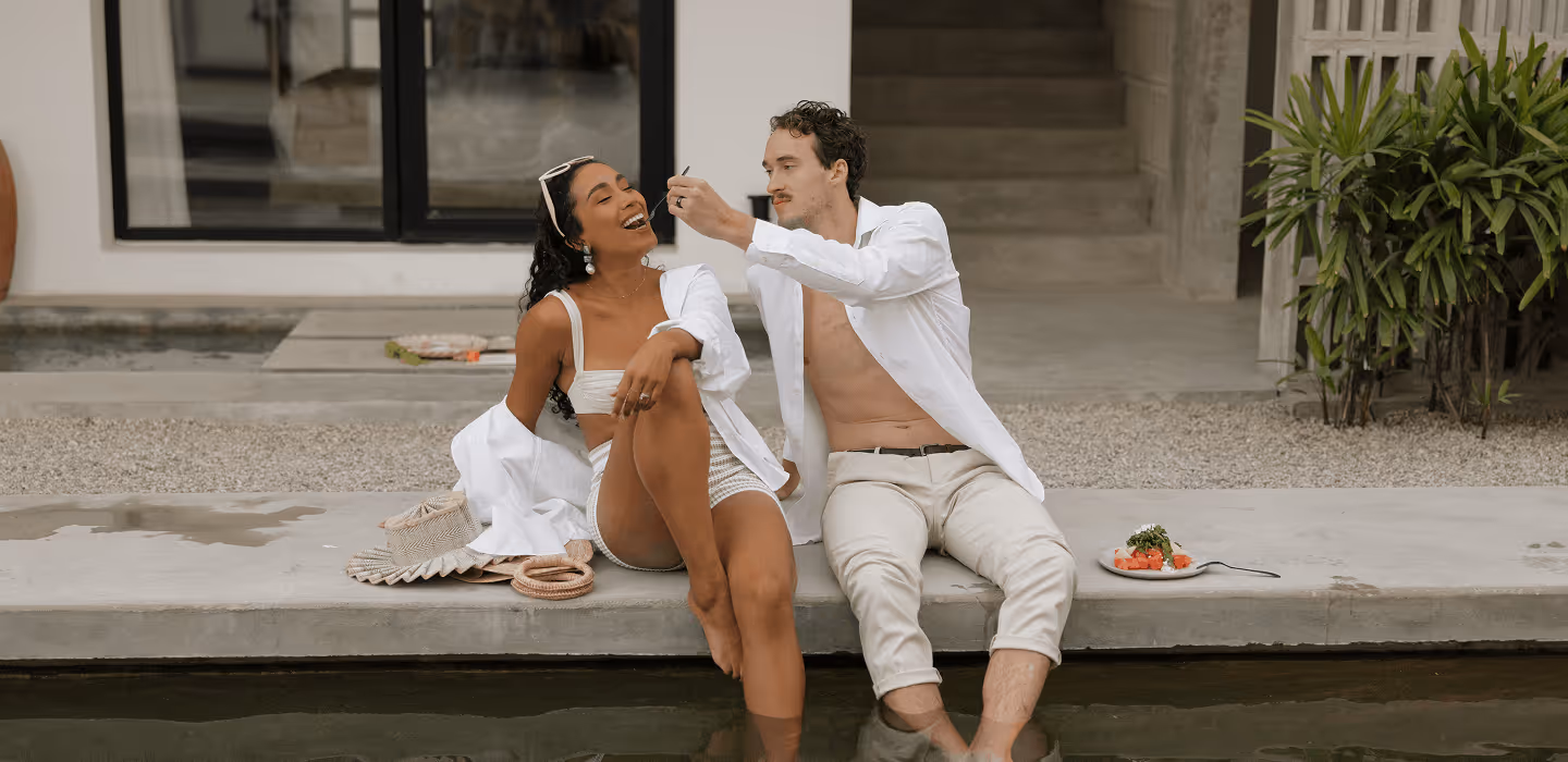Joyful couple sitting poolside as the man feeds the laughing woman, both dressed in casual white resort-style outfits