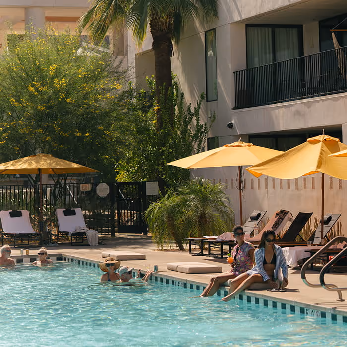 Hotel pool area with yellow umbrellas, lounge chairs, palm trees, and a guest walking poolside on a sunny day.