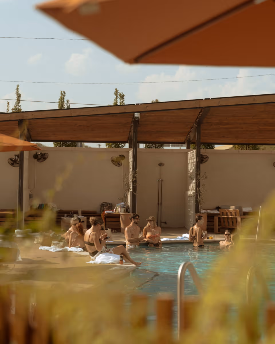 Friends relaxing poolside on a sunny day, sitting on the edge of a turquoise pool under a wooden pergola with orange