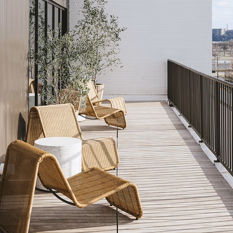 Sunny hotel balcony with woven rattan lounge chairs, a white terrazzo side table, and potted olive trees along a wooden deck.