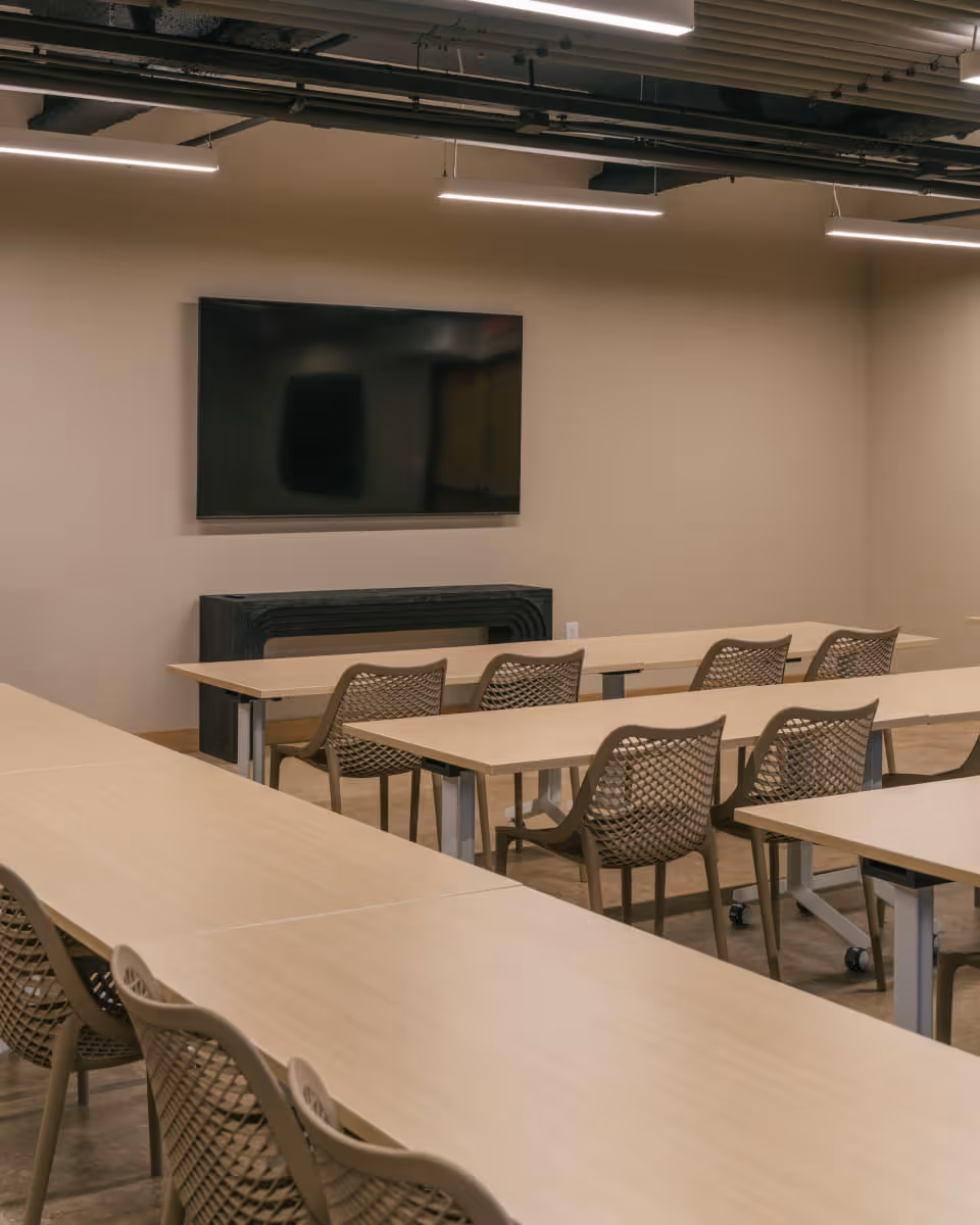Modern conference room with rows of light wood tables, mesh-back chairs, and a large wall-mounted screen