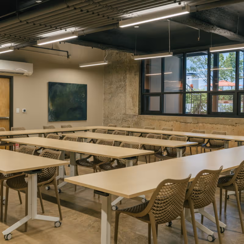 Spacious industrial meeting room with light wood folding tables, mesh chairs, concrete columns, large windows, and abstract