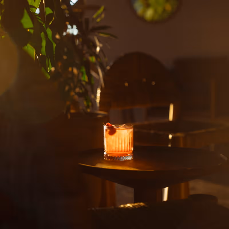 An amber-hued cocktail garnished with a cherry sits on a small round table in a dimly lit lounge with soft warm lighting.