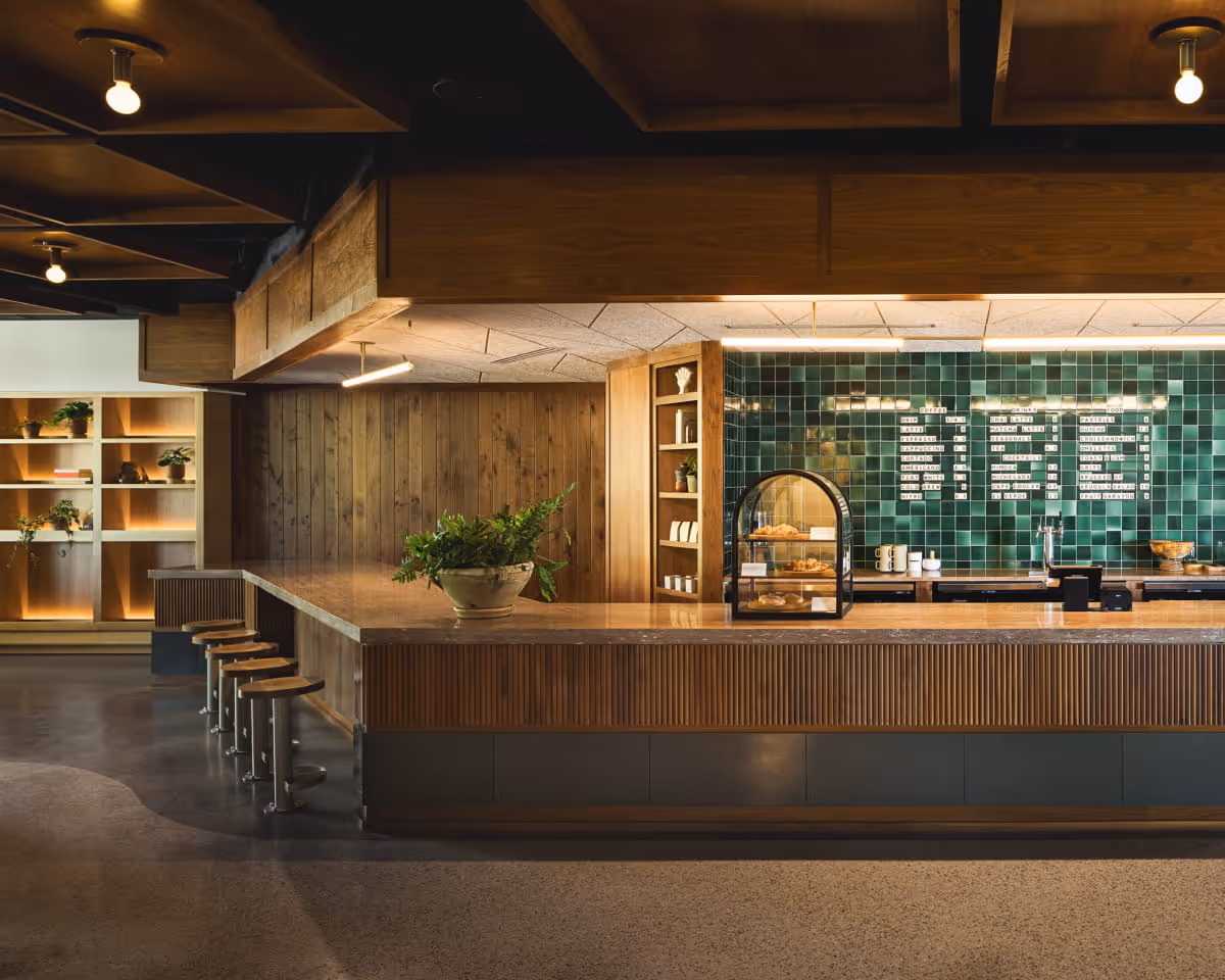 Modern café interior with warm wood paneling, fluted counter, green tiled backsplash displaying a menu, bar stools,