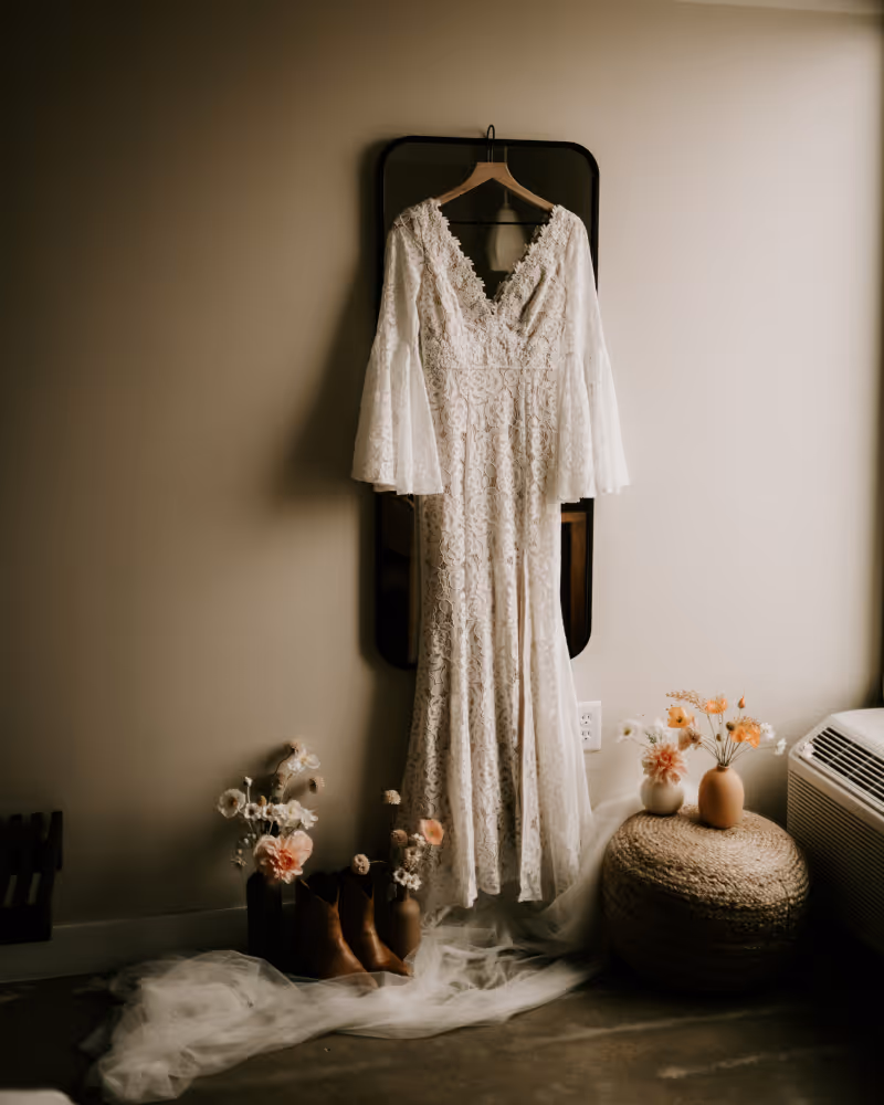 White lace bohemian wedding dress with bell sleeves hanging on a mirror, surrounded by floral arrangements and cowboy boots.