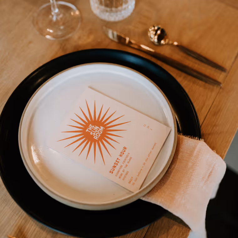 Place setting featuring The Sun Room Sunset Hour menu card on stacked black and white plates with a linen napkin.