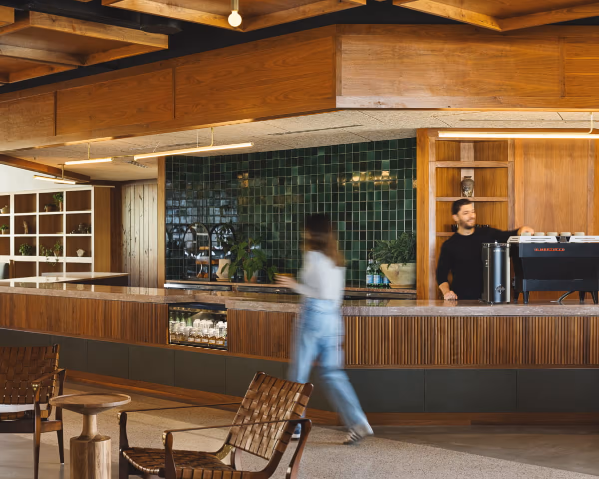 Warm wood-paneled coffee bar with green tile backsplash, espresso machine, woven leather rocking chair, and a barista