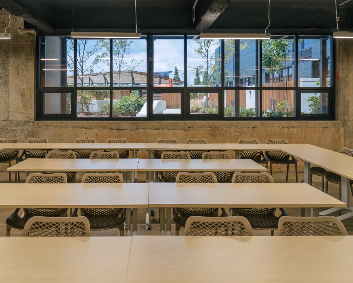 Modern classroom or meeting space with rows of light wood tables, mesh-back chairs, exposed concrete walls, and large