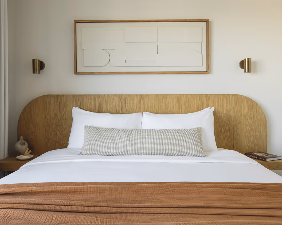 Modern hotel bed with a curved wooden headboard, white linens, a textured bolster pillow, and minimalist wall art above.