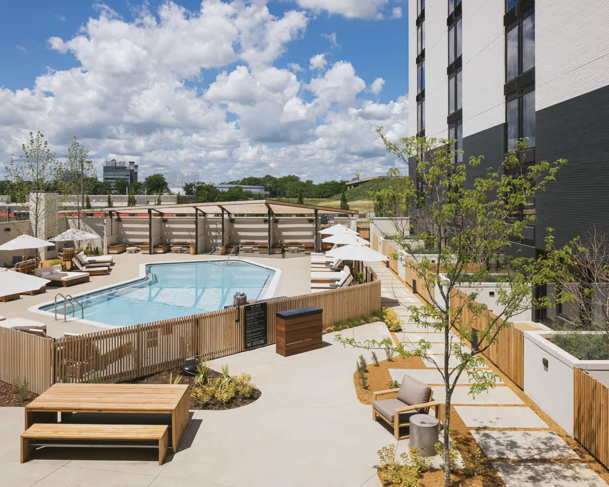 Outdoor pool deck with lounge chairs, white umbrellas, wooden fencing, landscaped pathways, and young trees beside a modern