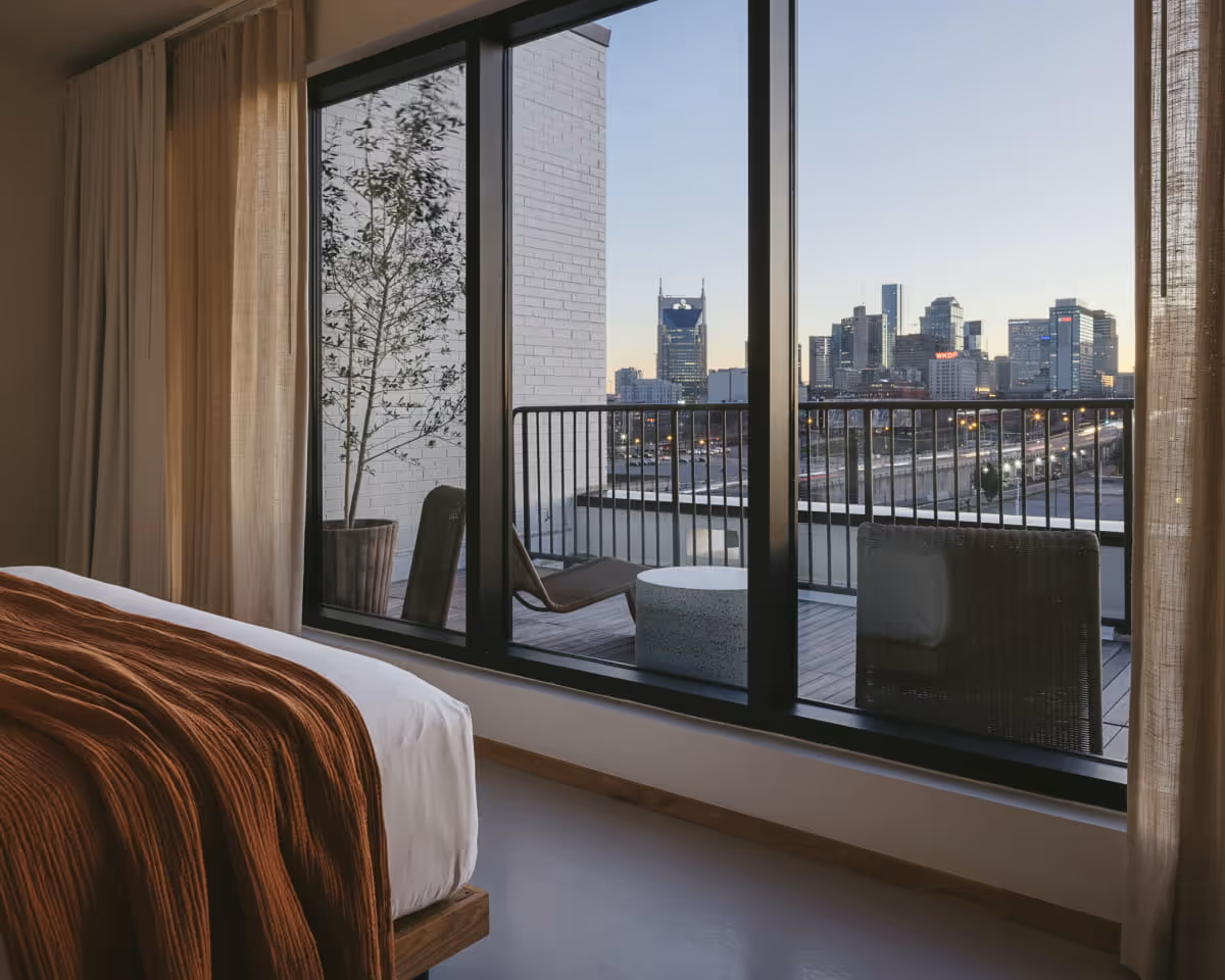 Hotel bedroom with a balcony overlooking the Nashville skyline at dusk, featuring modern outdoor furniture and sheer
