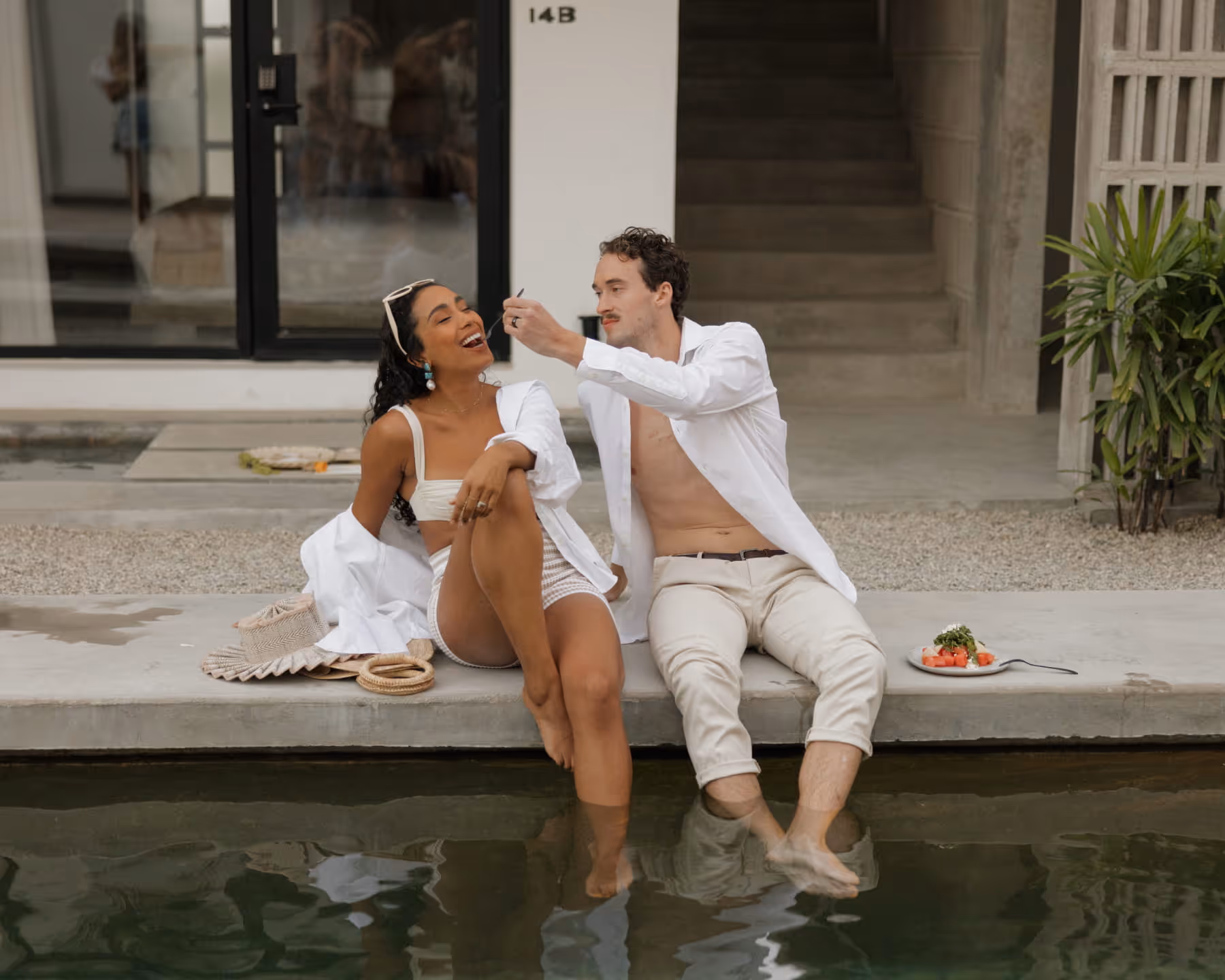 Joyful couple sitting poolside as the man feeds the laughing woman, both dressed in casual white resort-style outfits