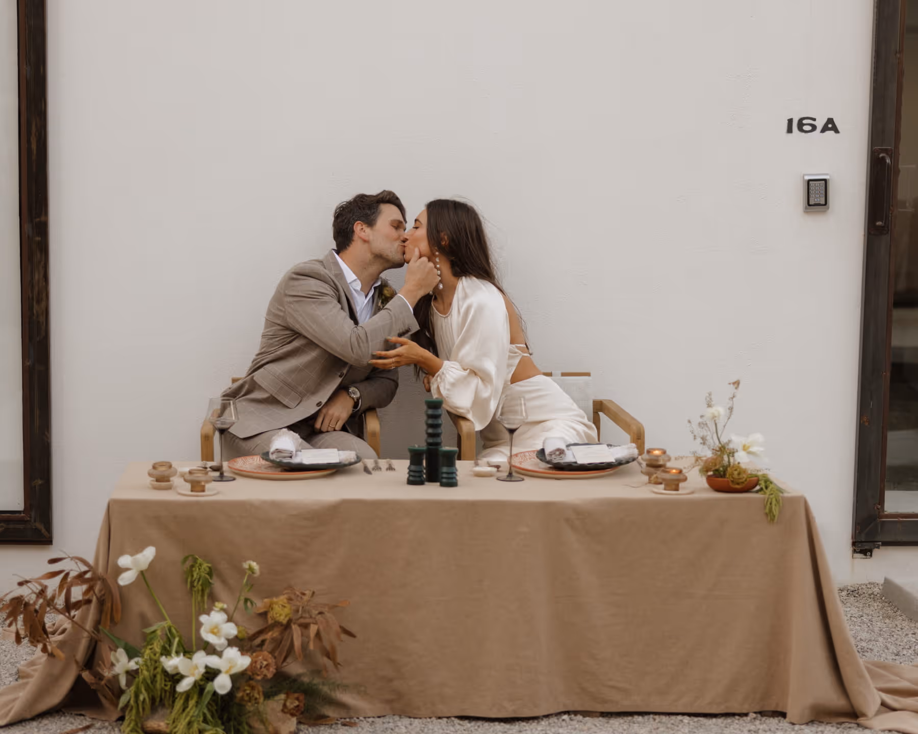 Newlywed couple sharing a kiss at their intimate sweetheart table decorated with earth-toned linens, candles, and ceramic