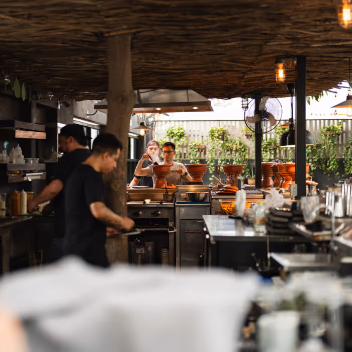 Bartenders working in a rustic open-air kitchen bar with a thatched roof, clay pots, and lush greenery in the background