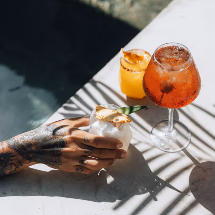 Tattooed hand holding a tropical coconut cocktail poolside alongside an Aperol spritz and a citrus drink in the sun