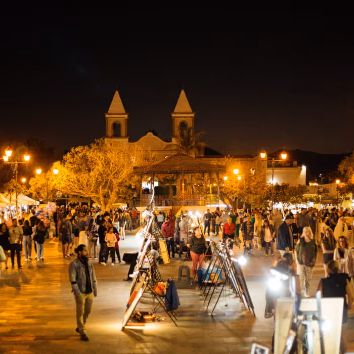Crowded nighttime plaza with art easels, a gazebo, and a twin-towered church illuminated by warm street lights in a Mexican