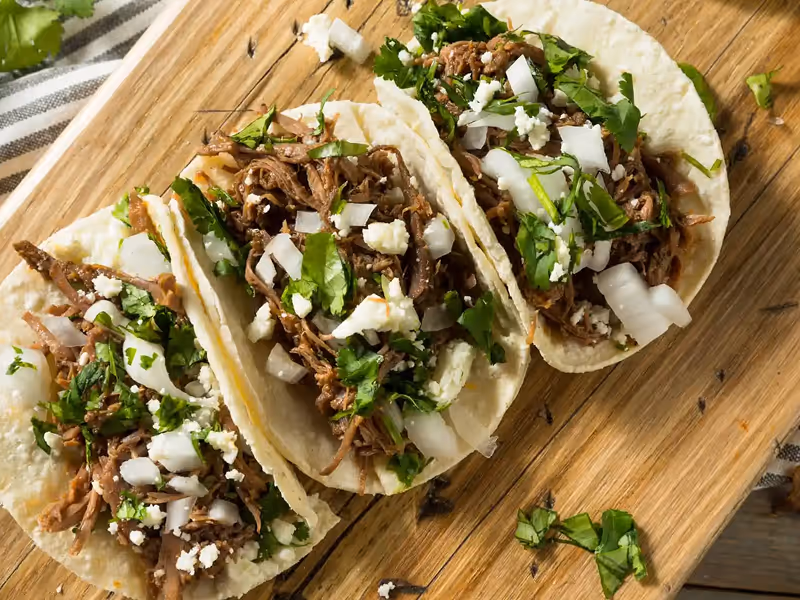 Hands holding fresh al pastor tacos topped with cilantro and onions at Taqueria El Paisa