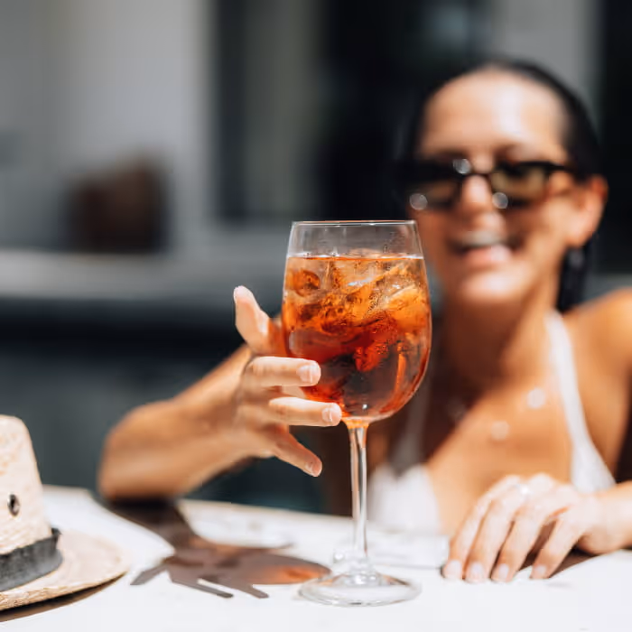 Smiling woman wearing sunglasses reaches for an Aperol Spritz cocktail in a wine glass on a sunny patio table