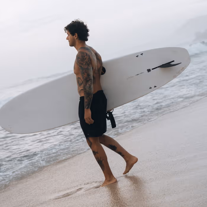 Tattooed surfer carrying a white surfboard under his arm while walking along a misty beach near the ocean waves
