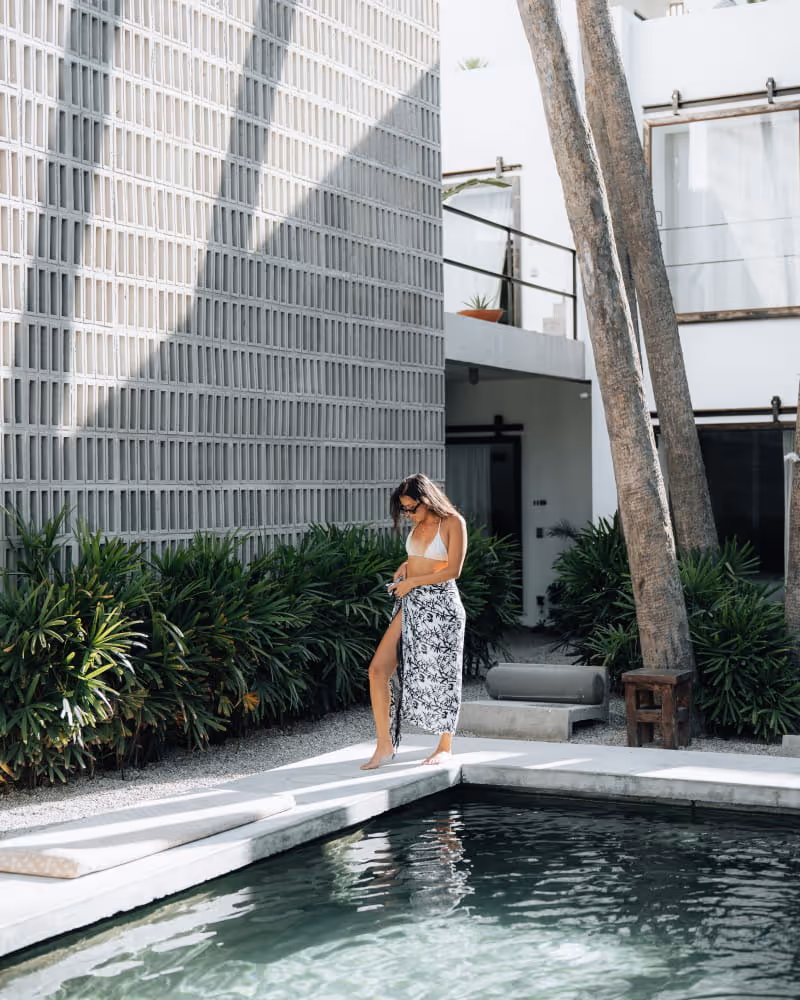 Woman in a white bikini and floral sarong walking barefoot along a modern boutique hotel pool with tropical plants
