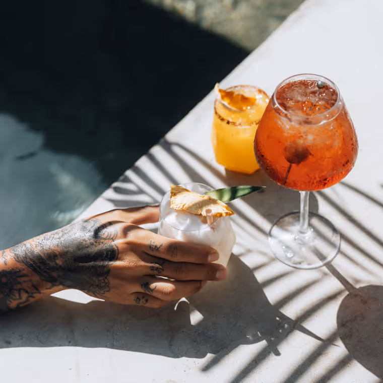 Tattooed hand holding a coconut cocktail beside an Aperol spritz and mango drink poolside in bright sunlight