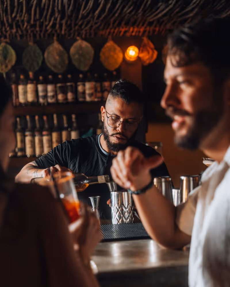 Bartender with glasses and tattoos pours a drink into a shaker while guests watch at a dimly lit thatched-roof bar