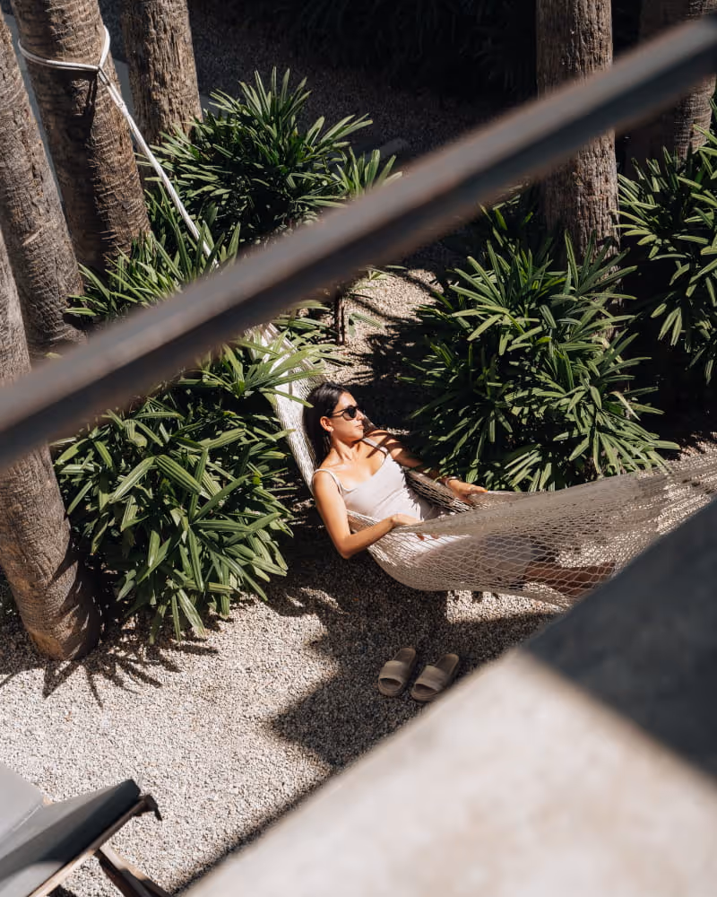 Woman relaxing in a hammock surrounded by tropical palm trees and lush greenery, viewed from a balcony above.