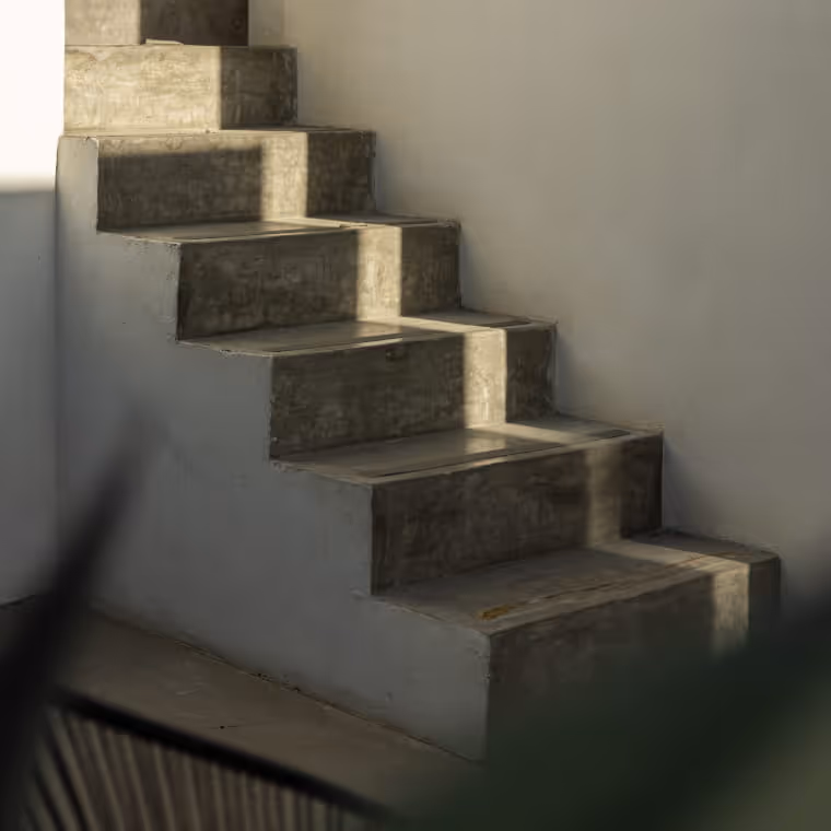 Sunlit concrete stairs ascending along a white plastered wall, casting geometric shadows in a minimalist architectural