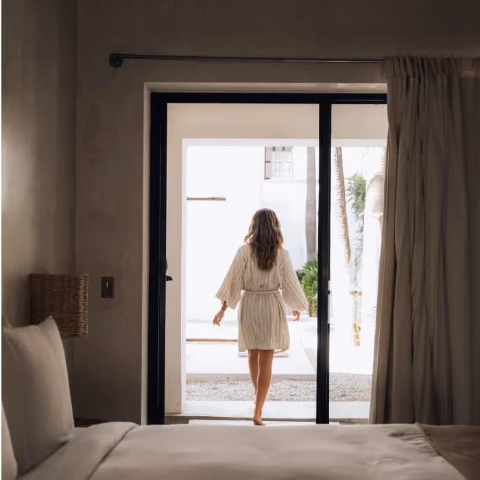 Woman in a linen robe walking barefoot through open glass doors from a moody bedroom into a bright sunlit courtyard.