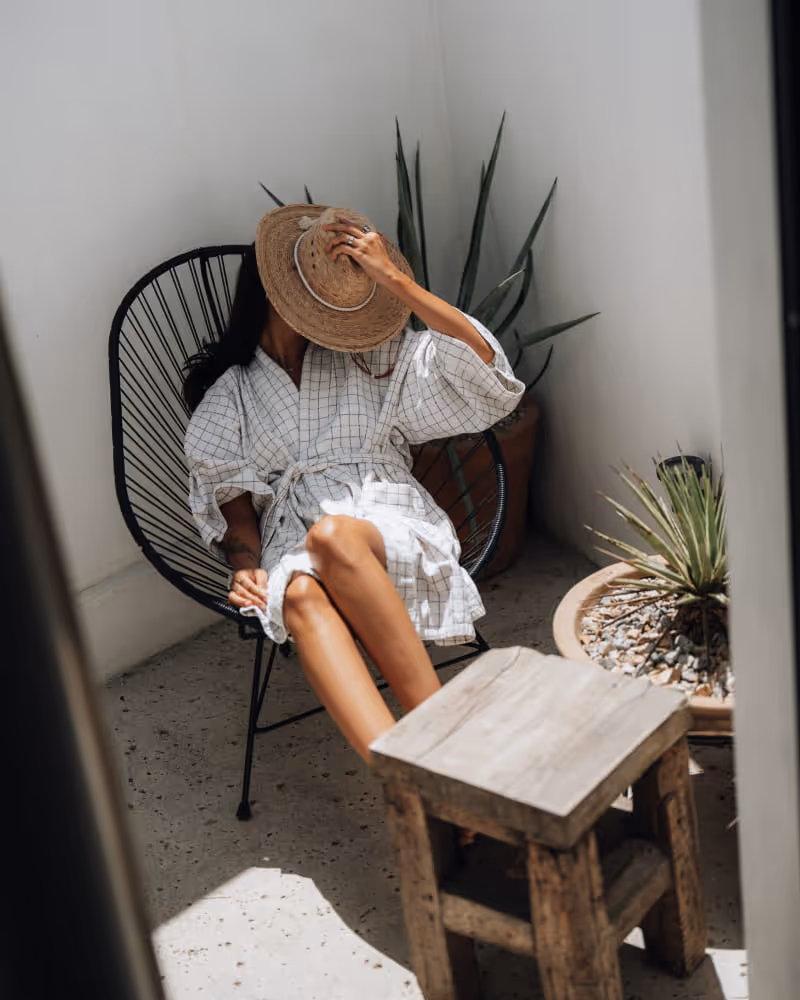 Woman in a linen dress and straw hat relaxing in a black Acapulco chair surrounded by desert plants and rustic decor