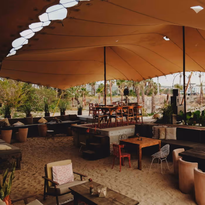 A bohemian beach lounge with a stretch tent canopy over sandy floors, eclectic furniture, and tropical palm trees