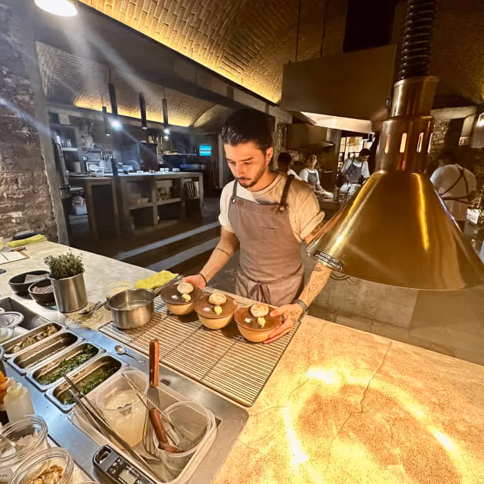 A chef in an apron plates desserts in clay bowls under warm pendant lights inside a professional open kitchen restaurant.