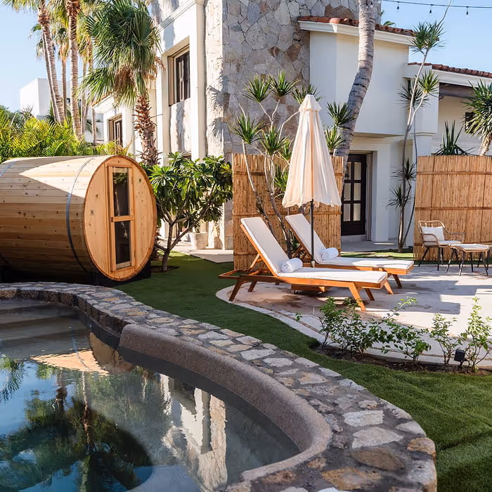 A resort spa area featuring a barrel sauna, cushioned lounge chairs, and a stone-edged plunge pool surrounded by tropical